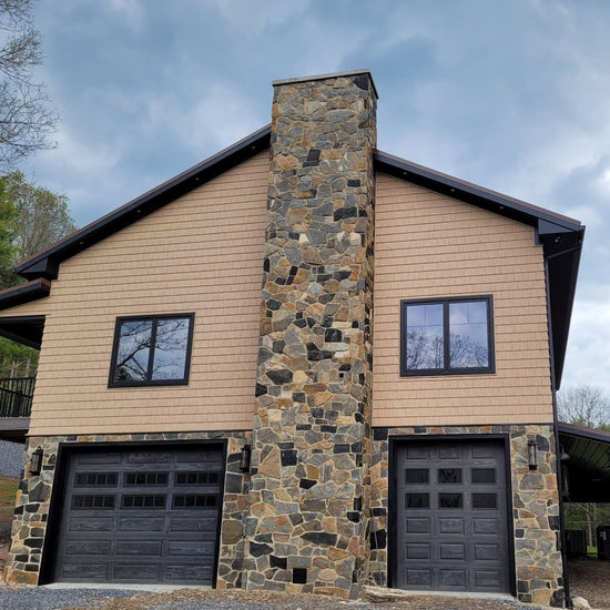 Two-story house with stone chimney and garage doors, surrounded by trees.